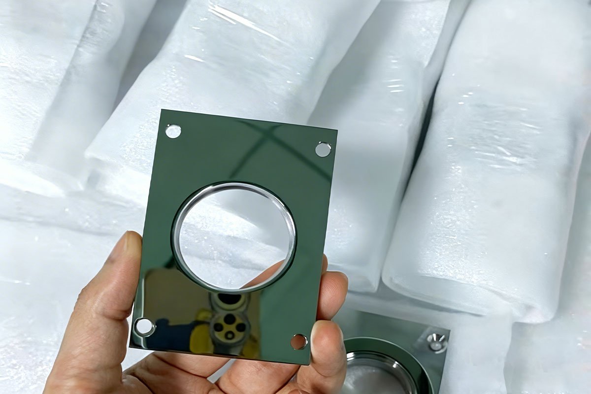 A custom high-gloss aluminum component, precision machined to a mirror-like finish, held against a background of protective foam packaging. The part features a central circular cutout and mounting holes, showcasing the smooth, reflective surface achievable through advanced CNC high-gloss cutting.