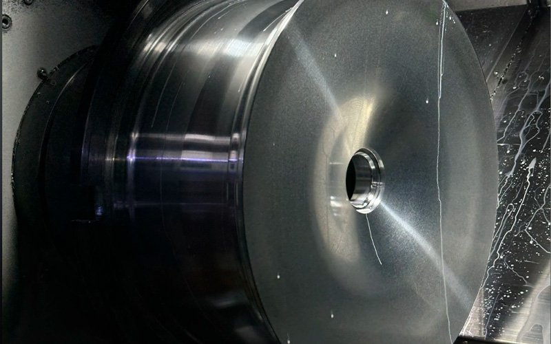 A solid forged aluminum wheel blank mounted on a CNC lathe, with the outer barrel and center bore freshly turned smooth, showing traces of machining coolant.