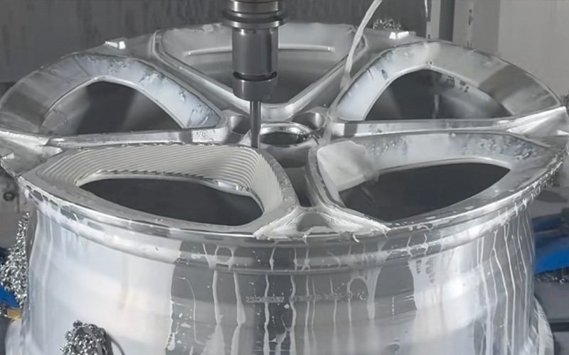A high-speed CNC milling spindle carving the intricate spoke design into a forged aluminum car wheel, with white coolant fluid flowing over the cutting area.