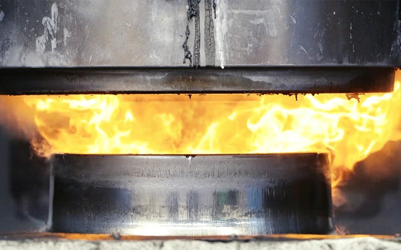 A heavy industrial press forging a superheated, glowing 6061 aluminum billet into a solid car wheel blank under extreme pressure.