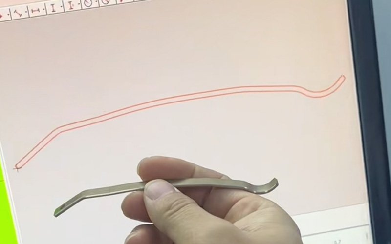 Hand holding a curved metal stamping part in front of a screen showing its measured 2D profile outline in red