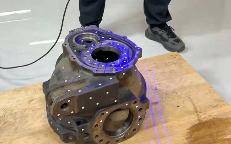 3D scanner projecting blue structured light onto a large industrial gearbox housing with white reference markers