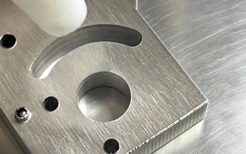 Dry ice nozzle deburring a CNC machined aluminum plate with an arc slot and counterbored holes, fine burrs visible along the milled edge