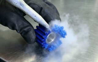 Dry ice blasting nozzle removing burrs from a blue machined plastic gear, with CO2 vapor sublimating on contact