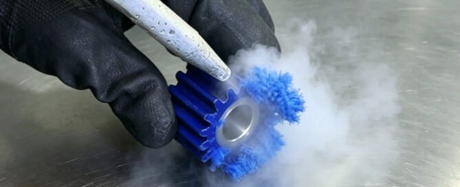 Dry ice blasting nozzle removing burrs from a blue machined plastic gear, with CO2 vapor sublimating on contact